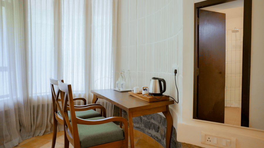A wooden desk set with 2 chairs, an electric kettle, and a tray near a tall window at Kadkani River Resort, Coorg.