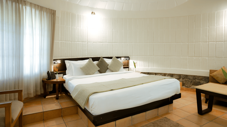 A cottage bedroom featuring a king size bed, side table, and curtained window at Kadkani River Resort, Coorg.