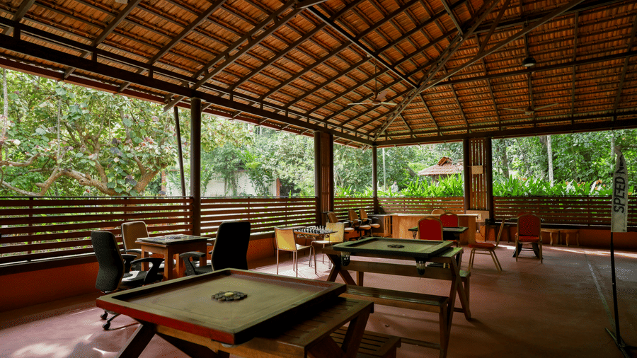 An open-sided recreational hall under a tiled roof, containing 3 carrom boards set on wooden tables with surrounding chairs, wood railings, and views of trees at Kadkani River Resort, Coorg.