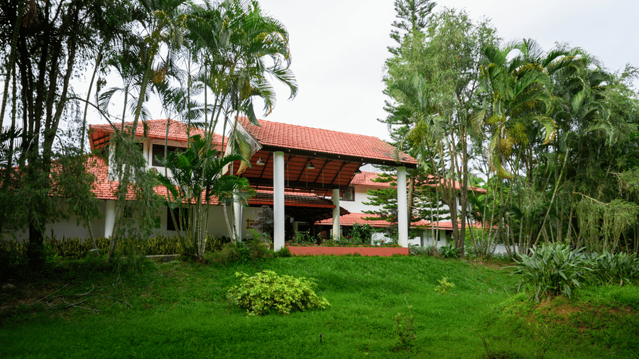 Front entrance porch with white pillars and red tiled roofs surrounded by green grass and tall trees at Kadkani River Resort, Coorg.