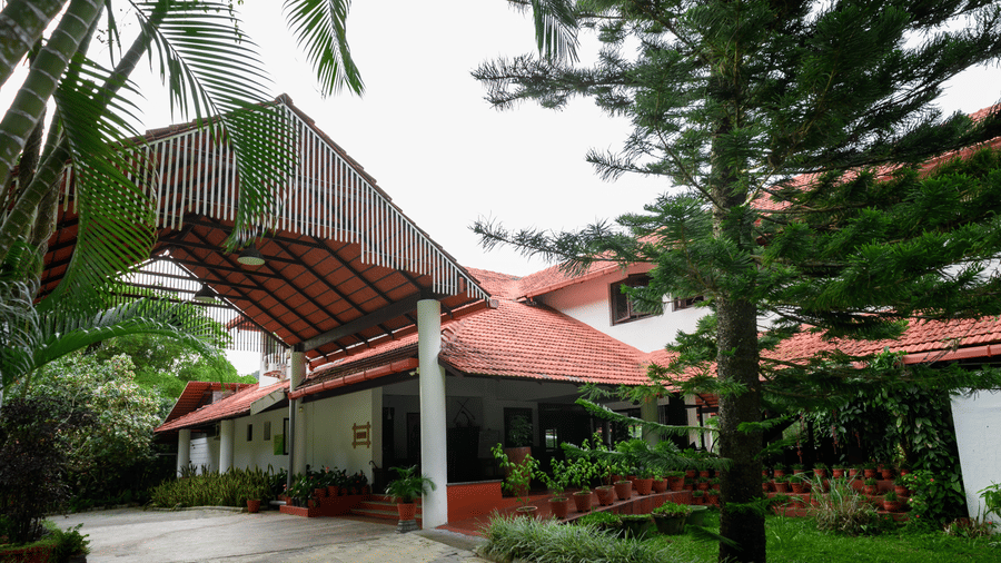 A resort building front with a covered gabled driveway entrance, roof tiles, white pillars, a tall pine tree, potted plants, and a paved driveway at Kadkani River Resort, Coorg.