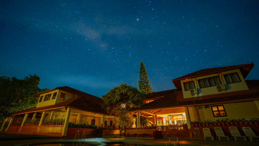 Buildings with illuminated windows and red tiled roofs next to a swimming pool under a dark starry sky at Kadkani River Resort, Coorg.
