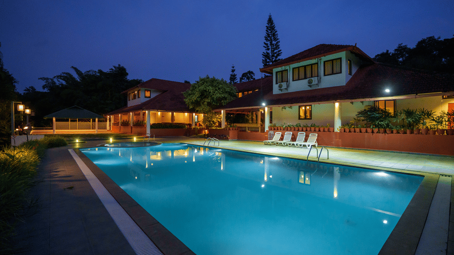 Rectangular swimming pool filled with water alongside resort buildings and sun loungers under a dark sky at Kadkani River Resort, Coorg.