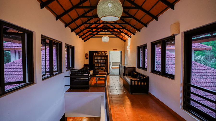 A high-ceilinged library hallway featuring bookshelves, open windows on both sides, wooden benches, tiled flooring, exposed roof rafters, and pendant lights at Kadkani River Resort, Coorg.
