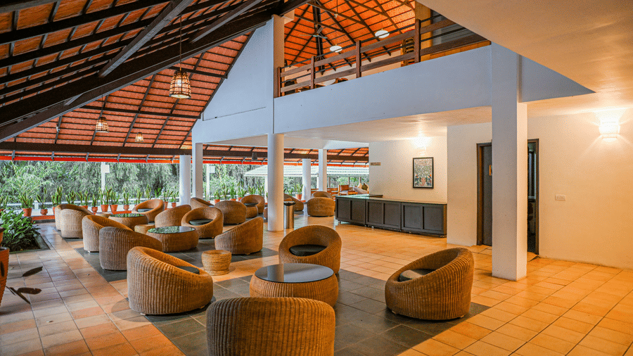 Open side lounge featuring tiled floors, sets of woven armchairs around round coffee tables, and white pillars at Kadkani River Resort, Coorg.