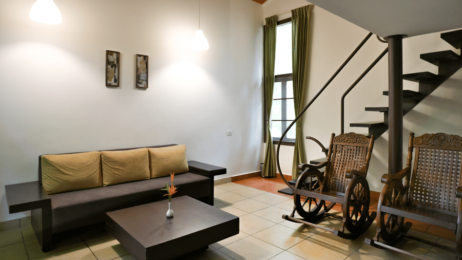 A living room at Kadkani River Resort, Coorg featuring a sofa set, square coffee table, 2 wooden rocking chairs, metal staircase and tall window with curtains.q