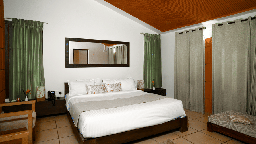 A bedroom at Kadkani River Resort, Coorg featuring a king size bed, wooden floor tiles, long mirror, curtains, side tables with lamps, small bench and seating area.