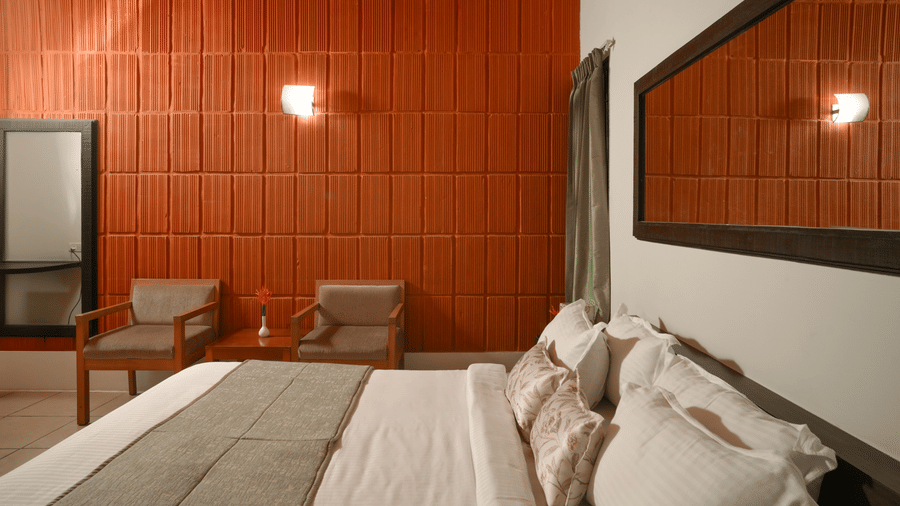A bedroom at Kadkani River Resort, Coorg featuring a king size bed with white pillows, wall mounted mirror, lamps, textured wall, wooden armchairs and center table.