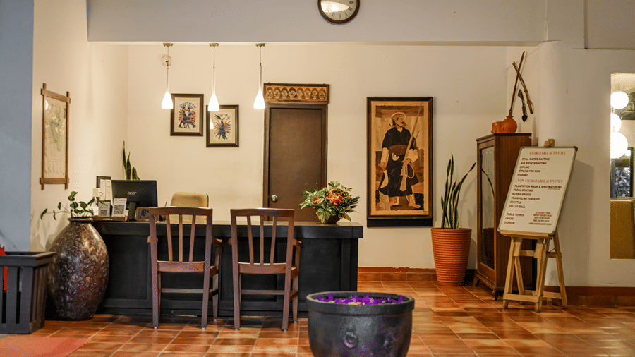 A dark wooden reception desk with 2 chairs, hanging lights, a large framed painting, potted plants, and terracotta floor tiles at Kadkani River Resort, Coorg.