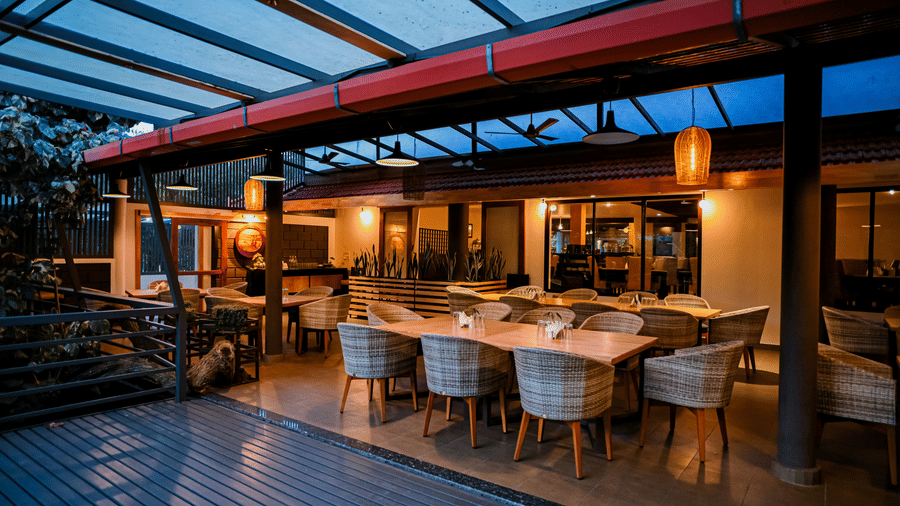 Dining area under a glass roof showing long wooden tables, woven chairs, hanging lanterns, and ceiling fans at Kadkani River Resort, Coorg.