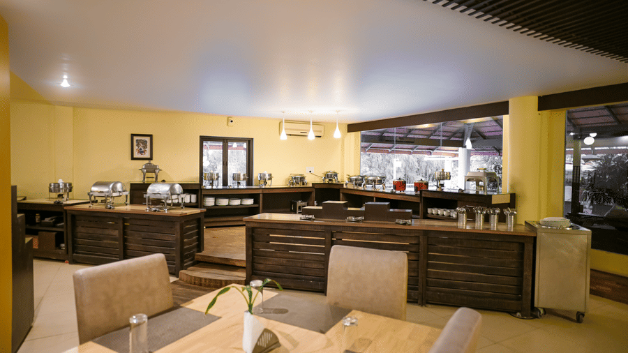 A buffet dining section with stainless steel chafing dishes on dark wooden counters, dining tables, place mats, and glass windows at Kadkani River Resort, Coorg.