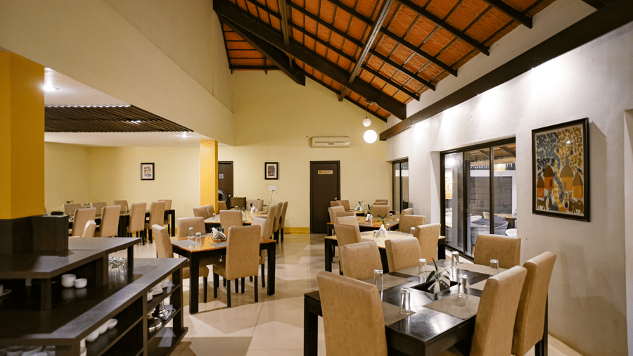 An indoor dining hall with a high sloped tiled ceiling, exposed dark wooden beams, wooden tables with beige chairs, and wall art at Kadkani River Resort, Coorg.