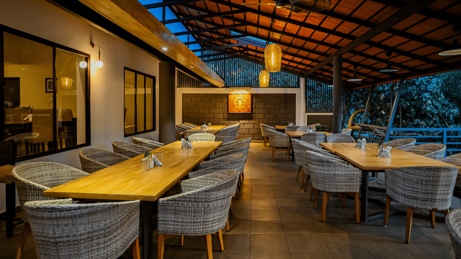 Semi outdoor dining space with long wooden tables, woven armchairs, hanging lights, and a wooden sloped ceiling at Kadkani River Resort, Coorg.