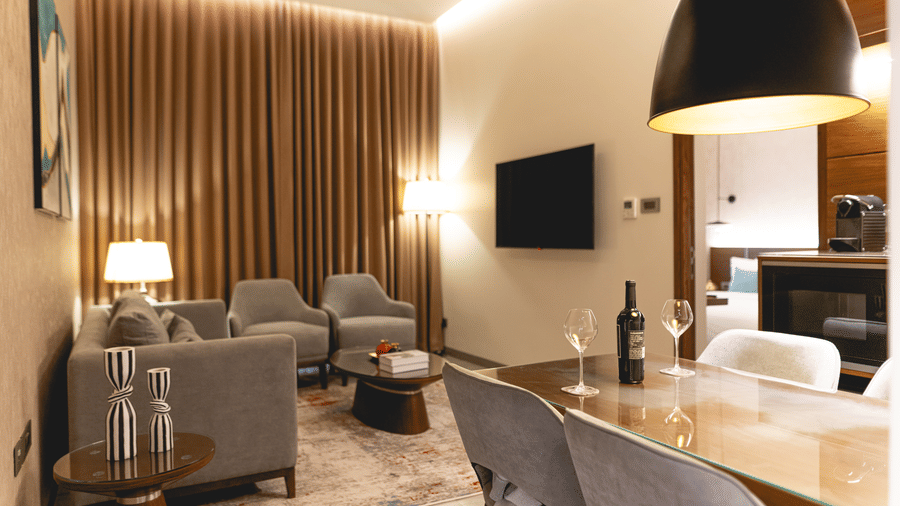 Modern living room at KertelSuites with neutral tones, featuring a sofa, armchairs, TV on the wall, floor and table lamps, and a dining table with chairs; warm lighting and vertical wooden slats on one wall.