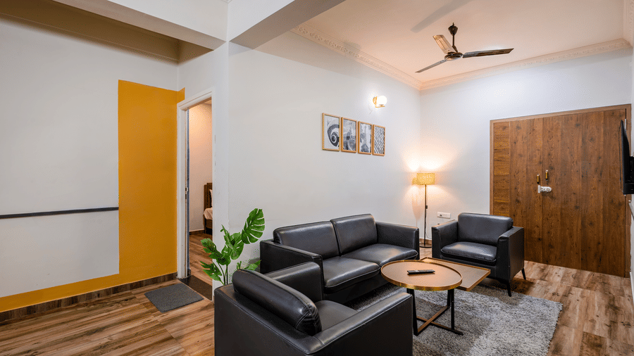 Diagonal view of the separate living room in Coral 4BHK of Kove Smart Stays, Hyderabad, seen with wooden flooring, upholstered multi-seating sofas, a ceiling fan, and a centre table.