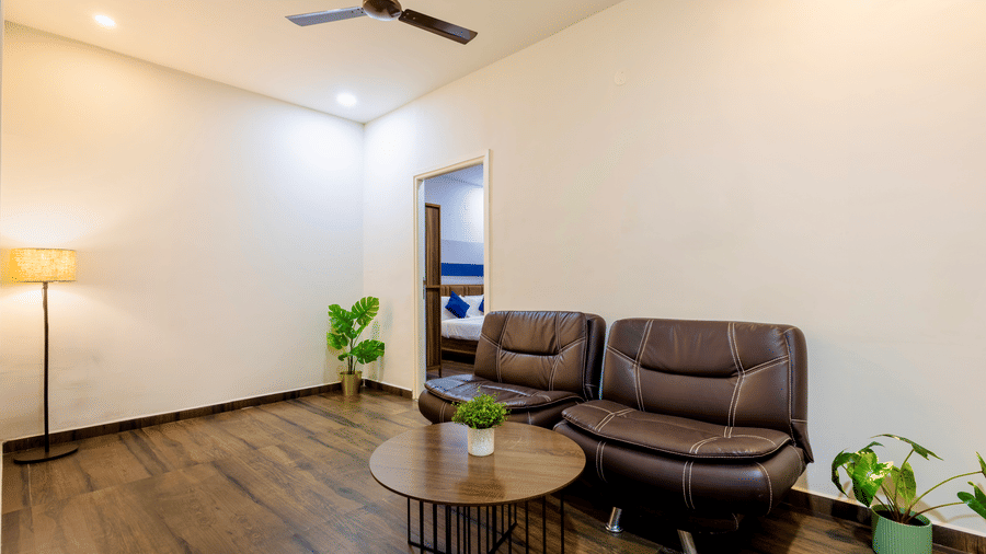 Spacious and separate living room in the Studio Room of Kove Smart Stays furnished with a multi-seater sofa with centre table, potted plants, and a lamp.