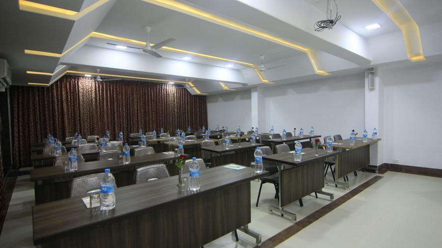 A well-lit conference room at Krushnai Resort with long rectangular tables arranged in class room style with chairs around them and a water bottle on each table.