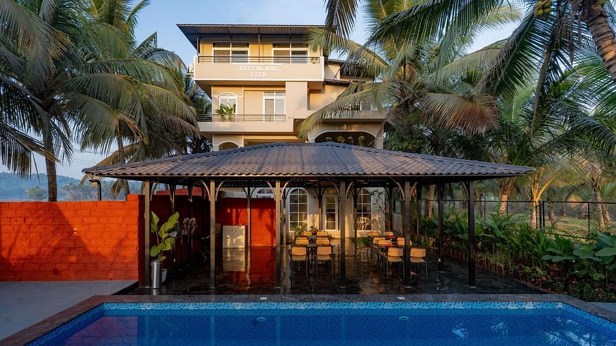 A view across a swimming pool towards a covered outdoor seating area with a thatched roof. Palm trees sway gently in the background at Kssemya.