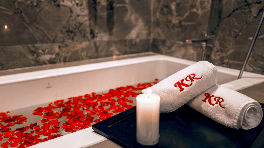 Close-up of a bathtub filled with water and floating petals, with a small candle and two rolled towels embroidered with the initials 'KR' at Kumar Resort by Turtle.