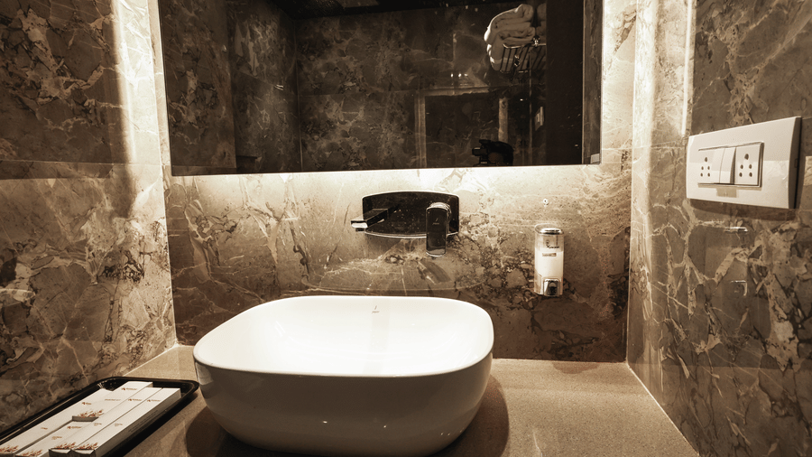 Close-up of a square basin on a vanity counter, set against a textured marble-effect wall, with a large mirror and electrical switches nearby at Kumar Resort by Turtle.