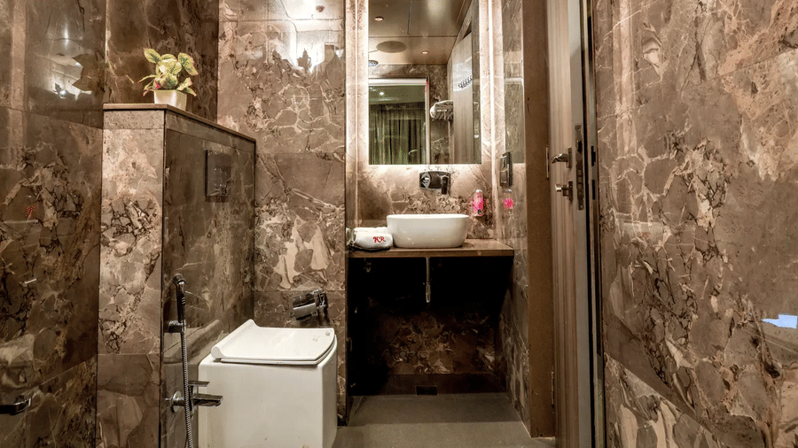Bathroom at Kumar Resort by Turtle with brown marble walls, vessel sink under back-lit mirror, wall-mounted toilet and a small potted plant on a shelf.
