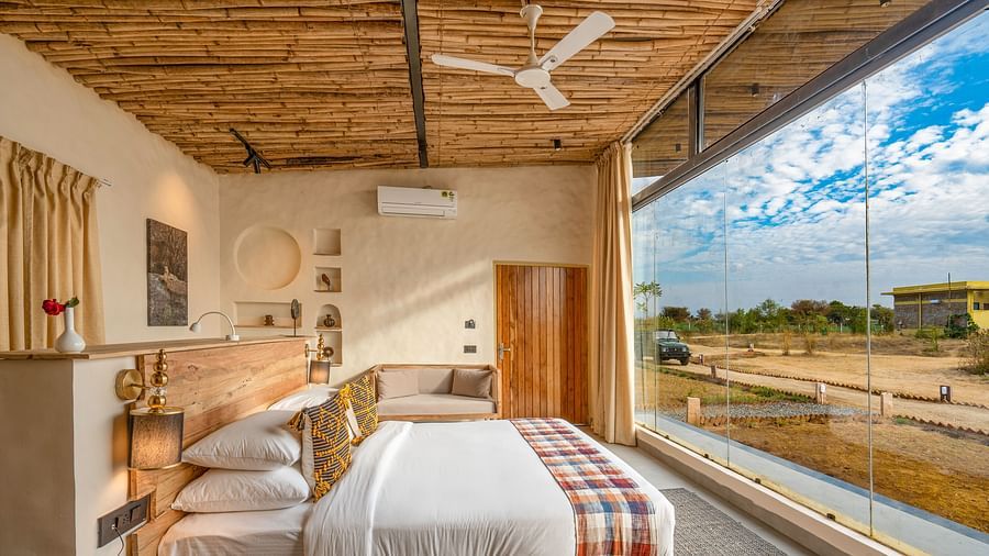 An image of a wooden furnished bedroom featuring a double size bed, a bedside table with lamp and socket, a sofa, some decorative elements in the room, and a large window from where outdoor scenery can be enjoyed - Kurja Jawai