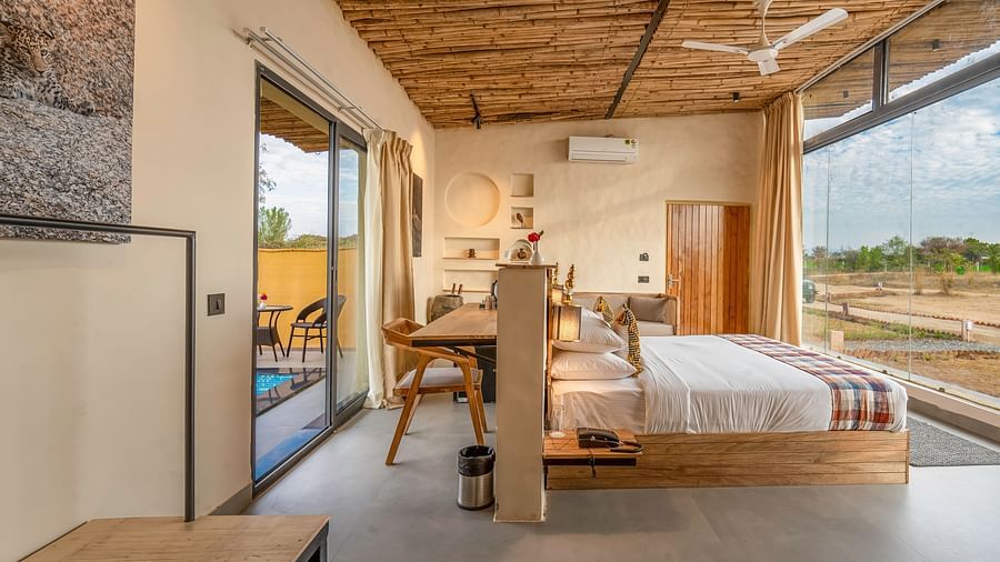 An image of Jungle Cottage bedroom featuring a bed, bedside table with lamp,  a work desk and chair behind, a sofa, AC, ceiling fan, and a large glass door at Kurja Jawai