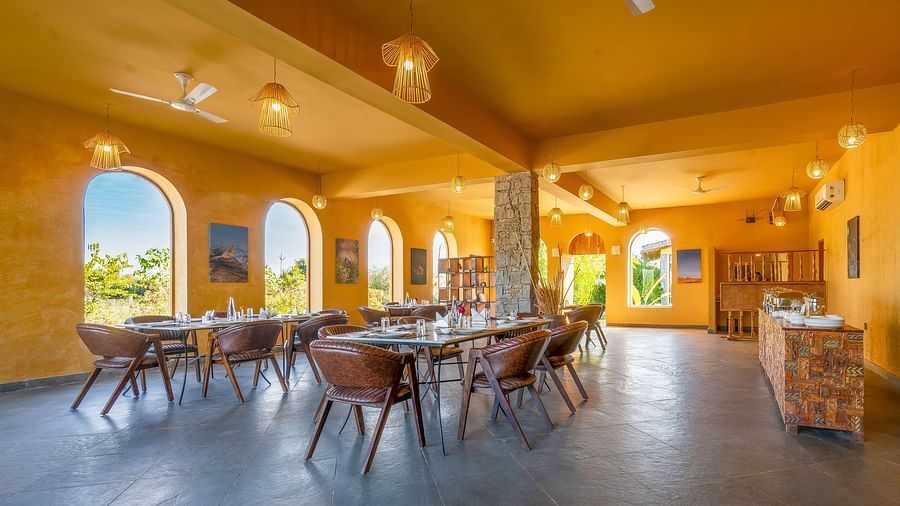 An image of L cave restaurant with boho set up featuring several sets of tables and chair,  lamp lights hanging from the ceiling, photo hanged on the walls and several open entrances - Kurja Jawai
