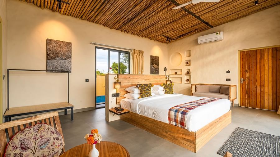An image of a wooden furnished bedroom  featuring a double size bed, a  chair and centre table, a sofa, AC, fan,  and some decorative items - Kurja Jawai