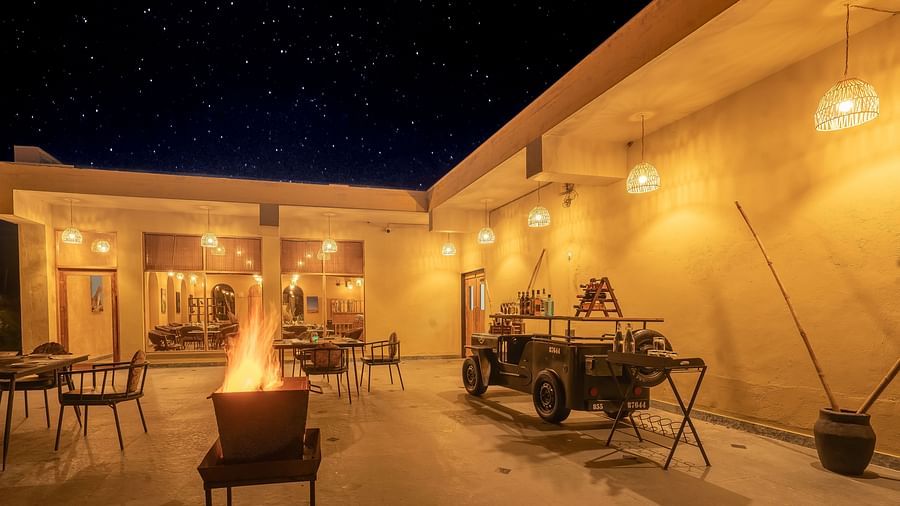 An image of open air Rosette Restaurant featuring several sets of table and chairs, a jeep without roof meant for decoration, and a pot where fire is buring at Kurja Jawai