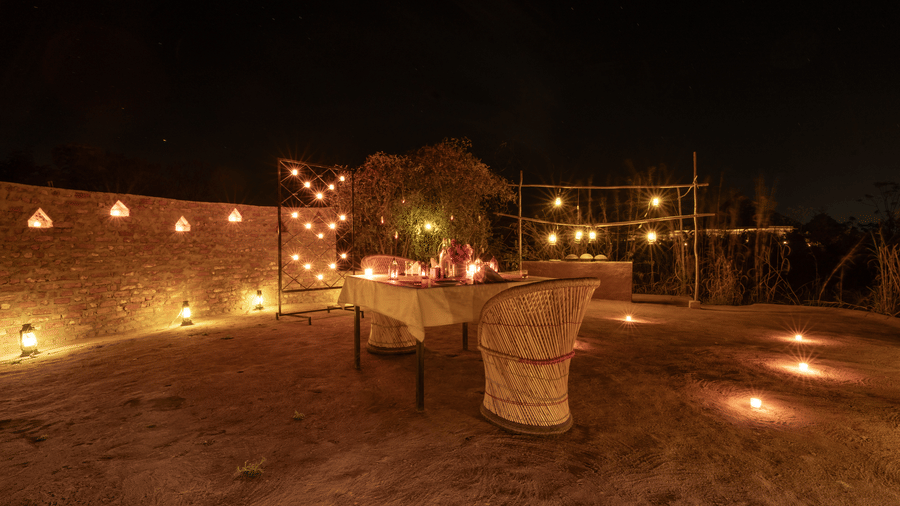 A warm, candlelit outdoor patio at night featuring a long table set for dinner with strings of glowing lanterns overhead.