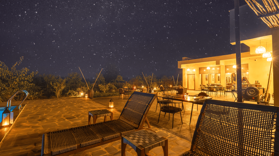 A swimming pool with a lit outdoor terrace at night featuring lounge chairs, overlooking a dark garden under a starry sky.