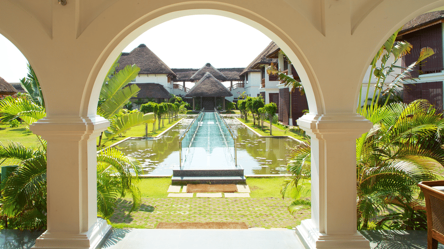 A view through a white arched opening towards a garden pool walkway and resort structures framed by palm leaves | Le Pondy