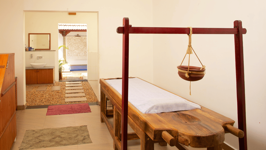 An Ayurvedic treatment room featuring a traditional wooden massage table setup with a hanging oil pot | Le Pondy
