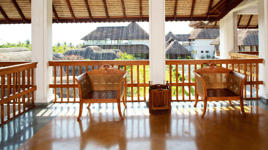 A covered outdoor balcony with a polished wooden floor, a wooden railing, and 2 wooden armchairs overlooking resort buildings | Le Pondy