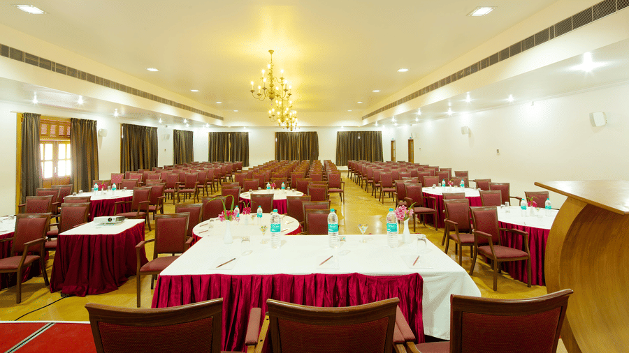 A large meeting room arranged with multiple tables covered in white tablecloths with red valances and surrounded by chairs | Le Pondy