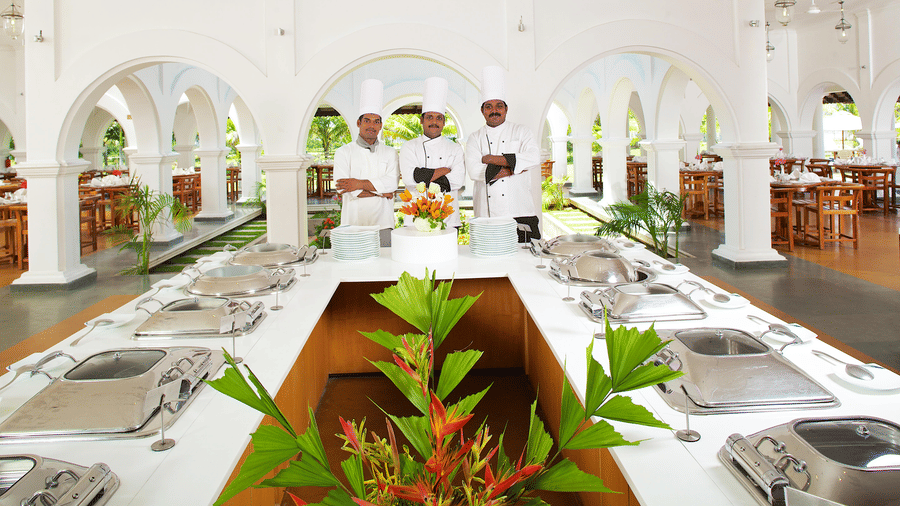 A buffet counter setup with stainless steel chafing dishes and 3 chefs standing behind it under arched columns | Le Pondy