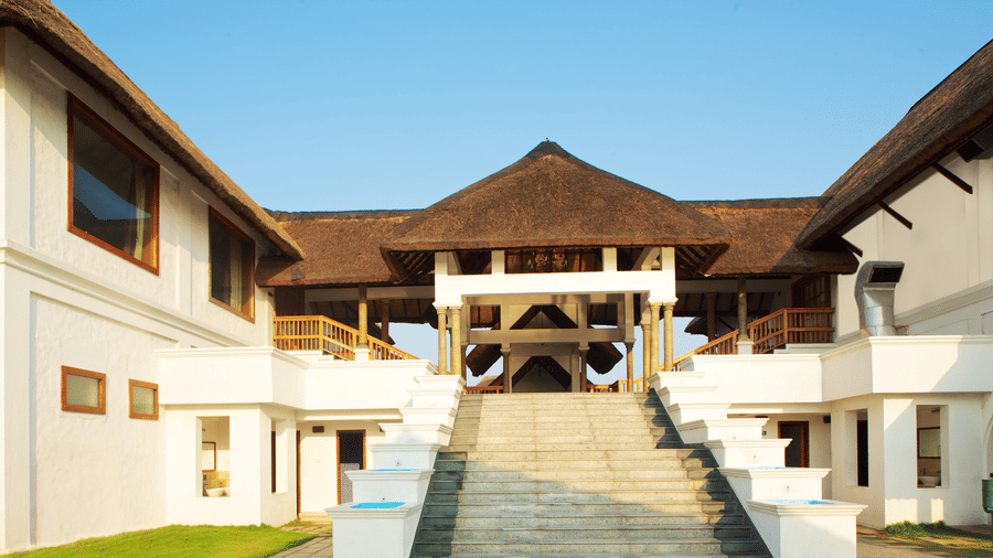 A stone staircase leading up to the main entrance of a large white resort building with a thatched roof | Le Pondy