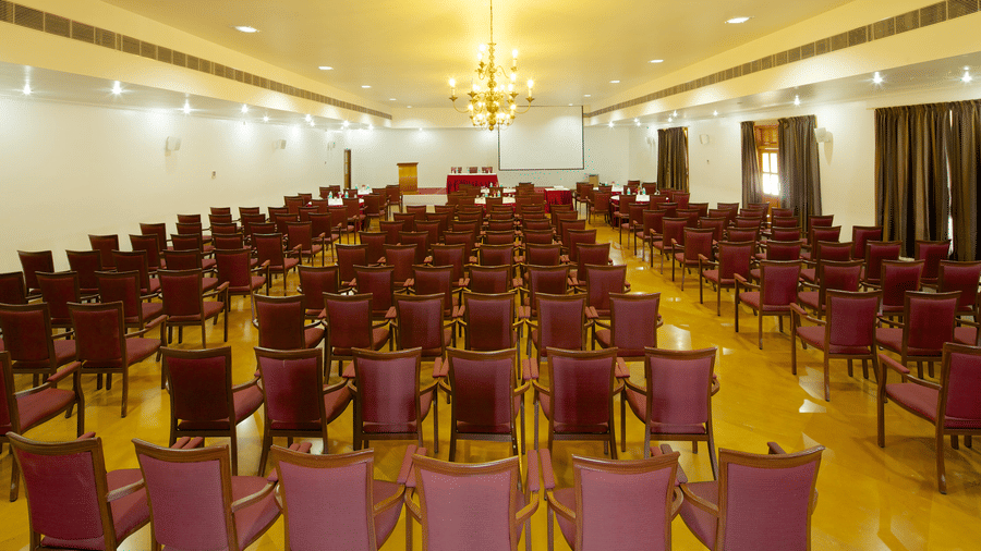 A large conference hall or auditorium with rows of wooden chairs with maroon upholstery facing a small stage under a chandelier | Le Pondy
