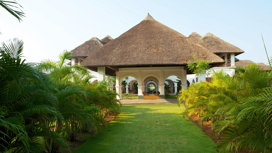 A manicured green lawn path lined with dense palm bushes leading towards a thatched-roof resort building | Le Pondy