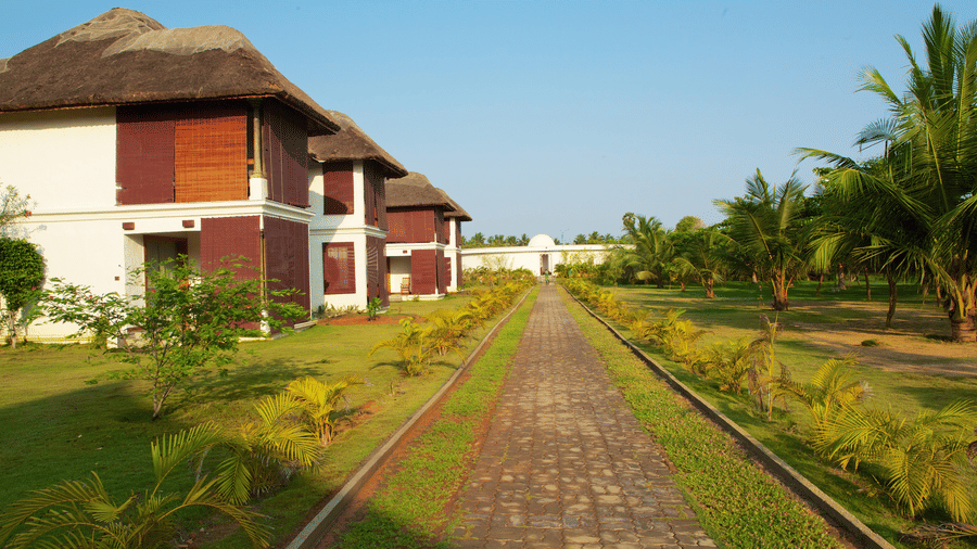 A stone-paved pathway running through a green lawn flanked by resort villas and young palm trees | Le Pondy