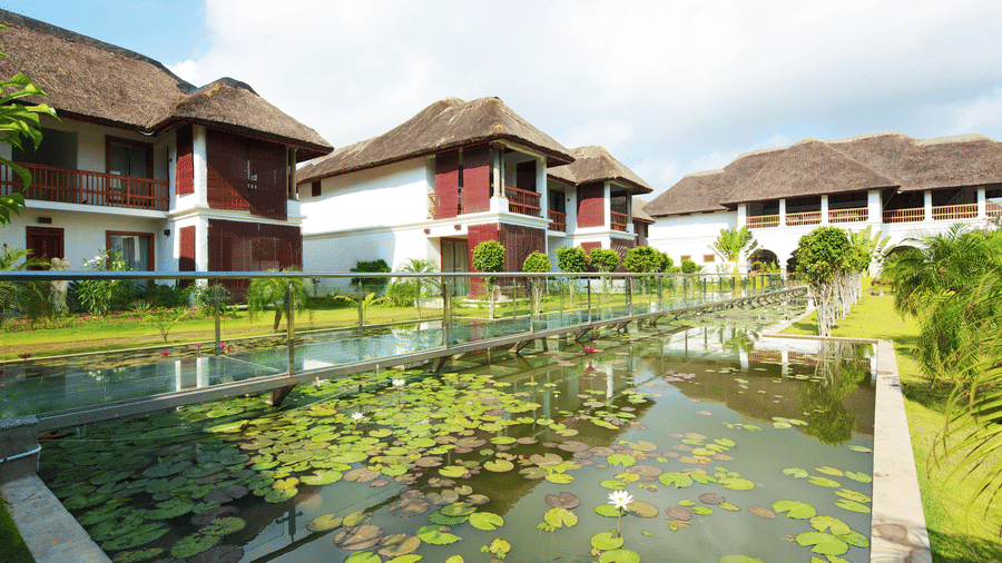 A transparent glass bridge with metal railings built over a pond covered with lily pads next to 2-storey resort buildings | Le Pondy