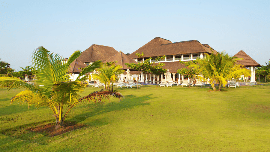 A wide green lawn with small palm trees situated in front of thatched-roof resort buildings | Le Pondy