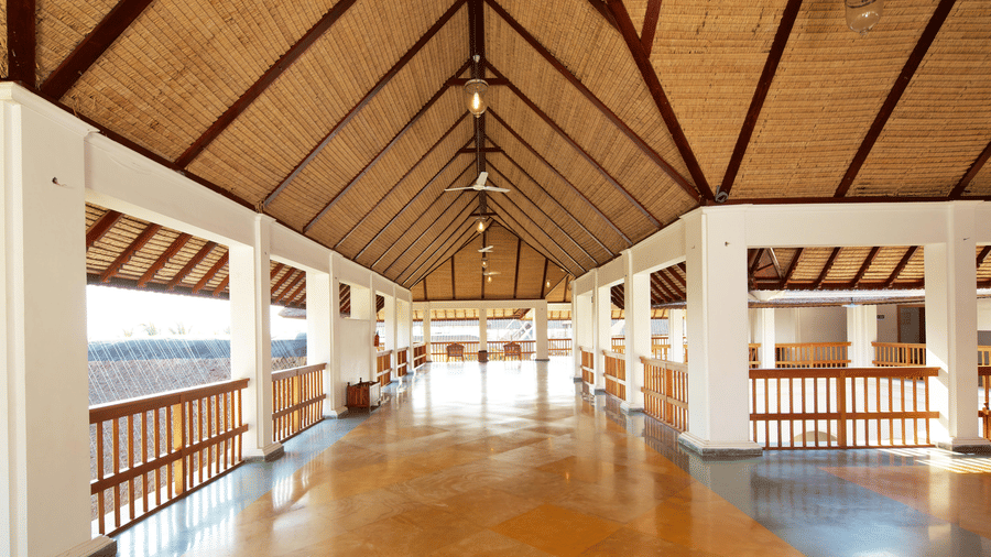 A wide, open-sided corridor with white pillars, wooden railings, a polished floor, and a high thatched-roof ceiling | Le Pondy