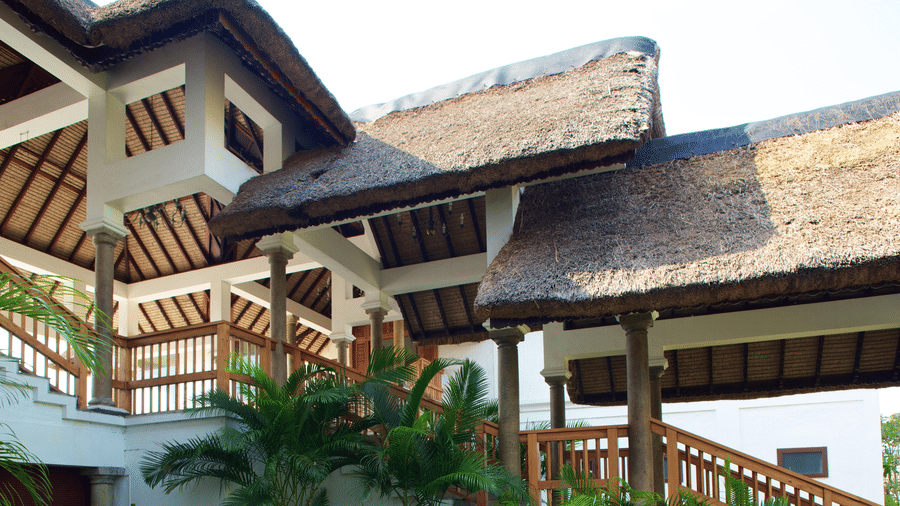 An exterior view of interconnected resort structures featuring white pillars, wooden railings, and thatched roofs | Le Pondy
