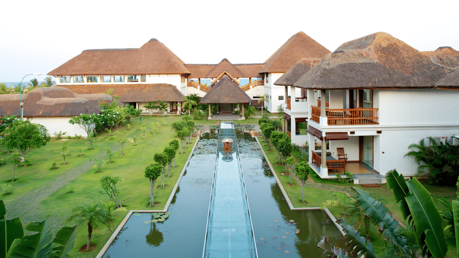 A multi-storey resort building with thatched roofs and balconies, featuring a long water body with a central glass walkway in the garden during daytime | Le Pondy