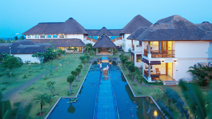 A multi-storey resort building with thatched roofs and balconies, featuring a long water body with a central glass walkway in the garden at dusk | Le Pondy