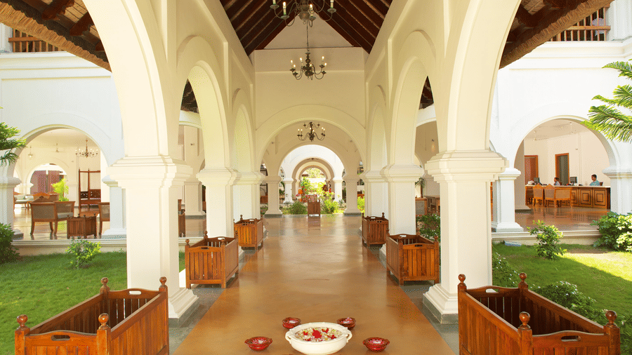 A spacious open-air resort lobby with white arched columns, wooden planters, and a floral decoration on the floor | Le Pondy