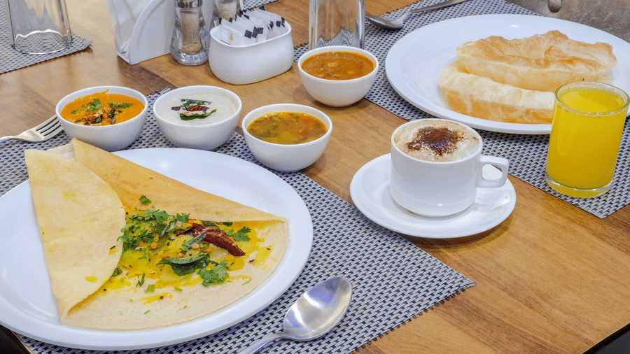 A South Indian breakfast spread at Lemonridge by Monday Hotels - Kukatpally features a filled dosa, small bowls of chutney, sambar, a coffee cup, and a glass of juice.