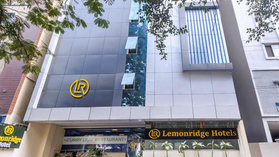 The modern exterior facade of Lemonridge by Monday Hotels - Kukatpally features building panels, window slats, potted plants, and tree branches.